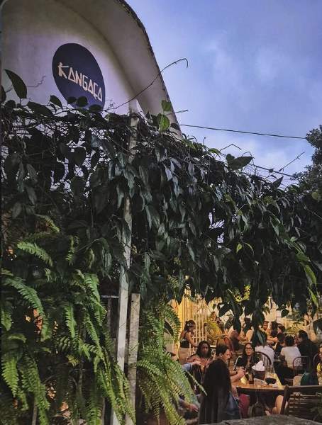 Fachada do Kangaça ao entardecer, com placa redonda azul e pessoas nas mesas sob as plantas