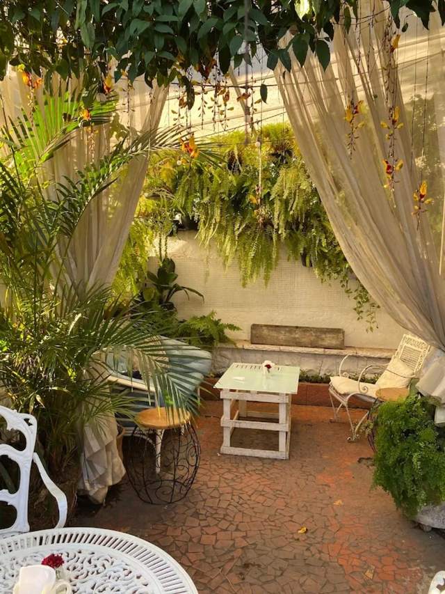 Jardim do Kangaça com cortinas claras abertas, samambaias e mesa branca sobre piso de cacos de cerâmica